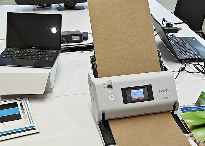 An A3 sheet-fed scanner in a laboratory feeding a board sheet from a batch, with a printed test sheet and a laptop on the table beside it.