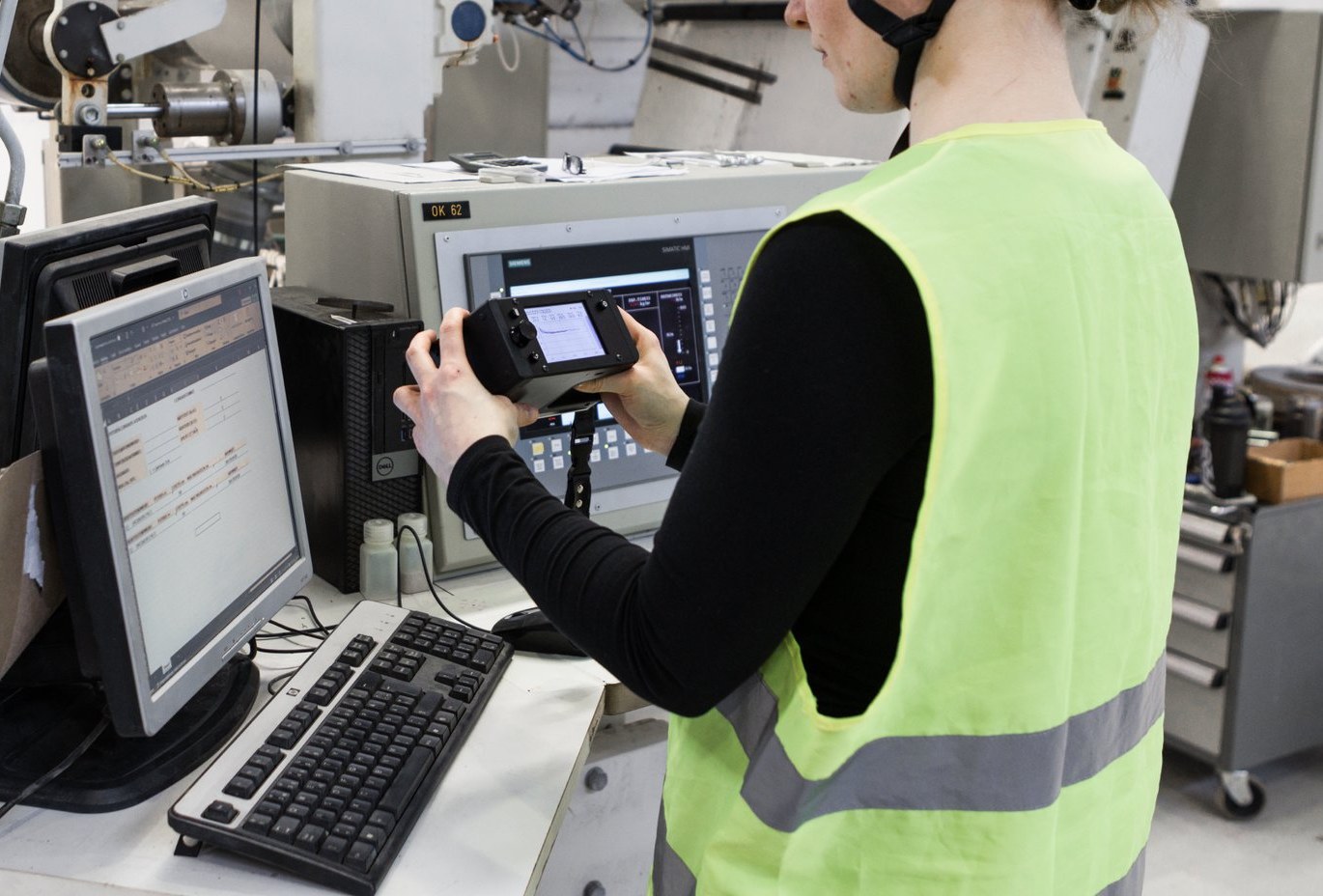 Person in high-visibility vest holding a Tapio RQP Live handheld device while working at a computer, with roll hardness data visible on both the device and a spreadsheet.
