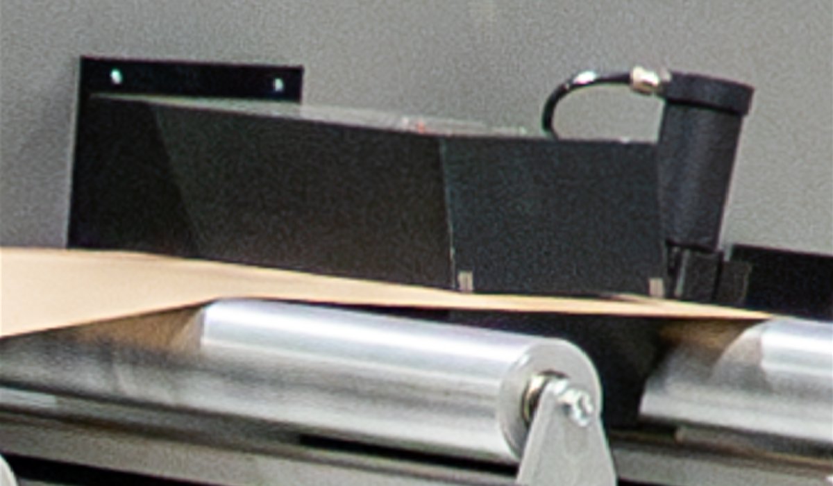 A close-up of the Tapio basis weight sensor positioned above a moving sheet on a roller bed, used for measuring grammage.