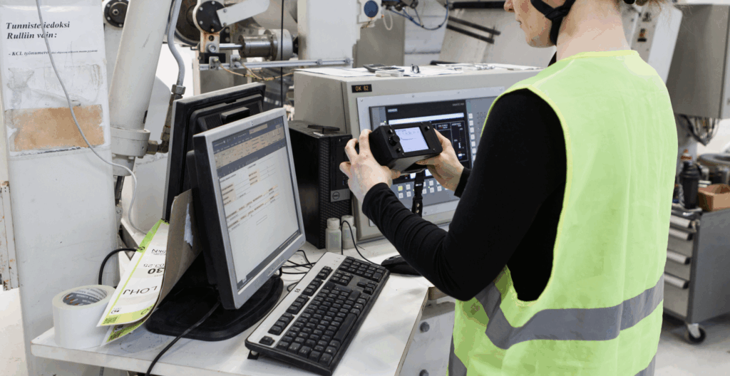 Person in high-visibility vest holding a Tapio RQP Live handheld device while working at a computer, with roll hardness data visible on both the device and a spreadsheet.