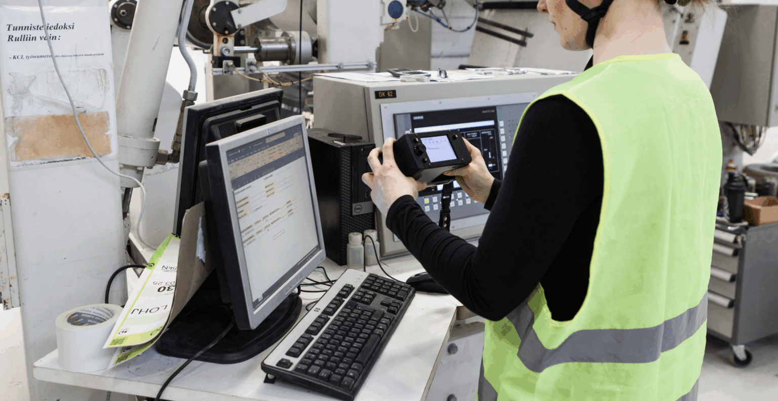 Person in high-visibility vest holding a Tapio RQP Live handheld device while working at a computer, with roll hardness data visible on both the device and a spreadsheet.