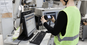 Person in high-visibility vest holding a Tapio RQP Live handheld device while working at a computer, with roll hardness data visible on both the device and a spreadsheet.