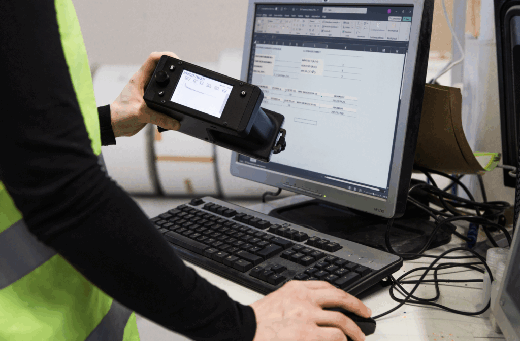 Person in high-visibility vest holding a Tapio RQP Live handheld device while working at a computer, with roll hardness data visible on both the device and a spreadsheet.