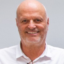 Smiling middle-aged man with a bald head and short beard, wearing a white shirt, photographed against a neutral background.