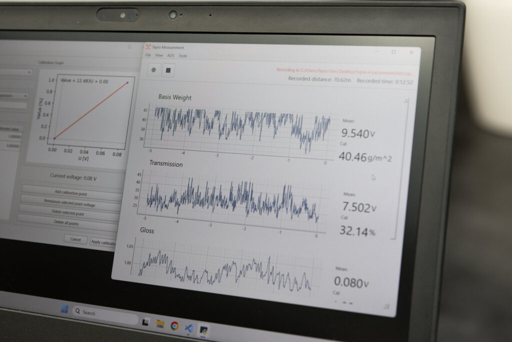 Laptop screen showing Tapio Measurement software during a paper measurement. The software displays live graphs for basis weight, transmission, and gloss, together with calculated measurement values.
