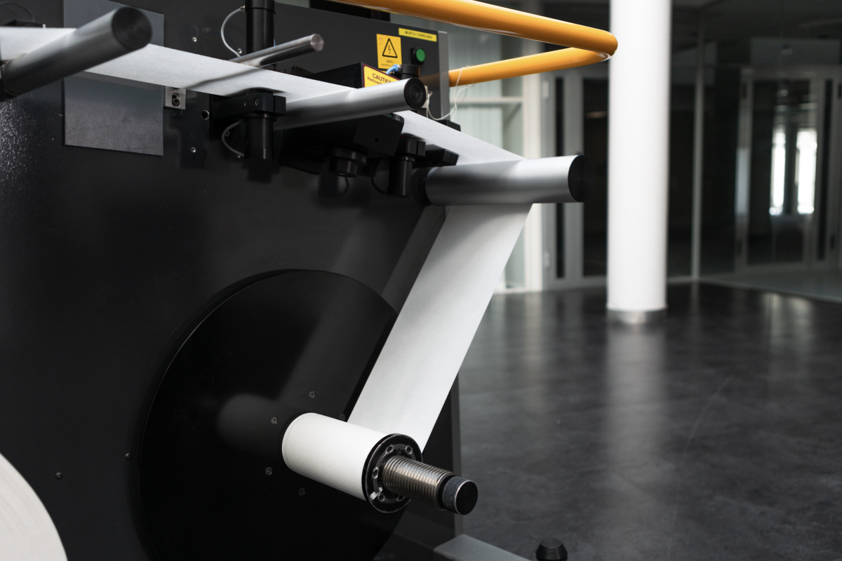 A laboratory paper testing device with a paper roll unwinding through a series of rollers and sensors enclosed in a sturdy frame.