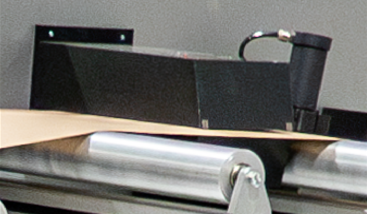 A close-up of the Tapio basis weight sensor positioned above a moving sheet on a roller bed, used for measuring grammage.